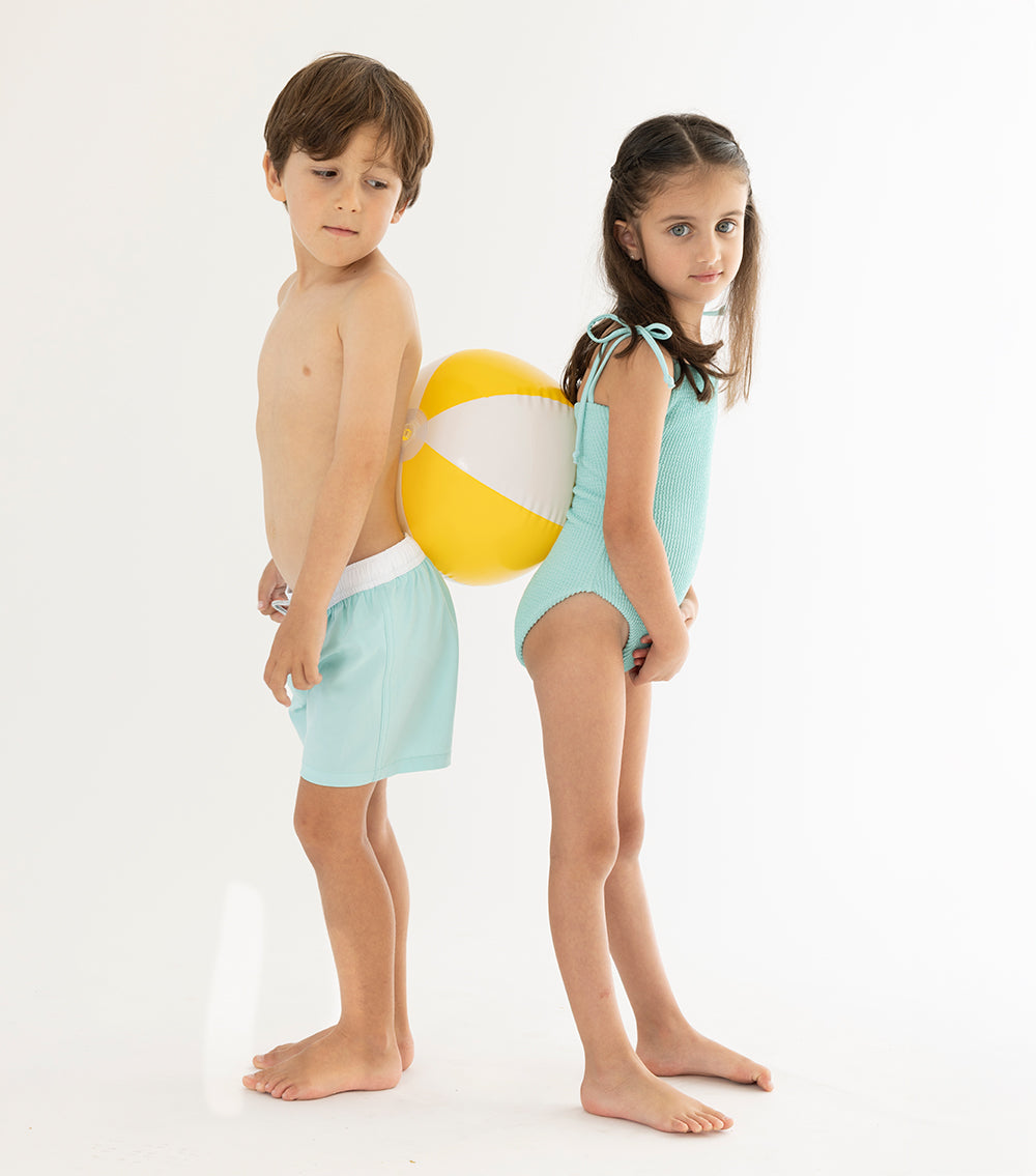 Aqua Two children in light blue swimsuits standing next to a beach ball on a white background