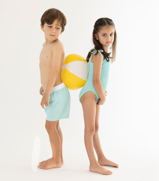 Aqua Two children in light blue swimsuits standing next to a beach ball on a white background